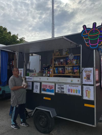 The image depicts a food stand in Sarandí, Argentina. The stand, with a distinctive neon sign, offers snacks and drinks. A man stands in front of the stand holding a small dog. Signage on the stand lists items such as "Fernet con Coca," daiquiris, and granizados with prices. The stand also displays a variety of chips and other snacks. The sky is overcast.