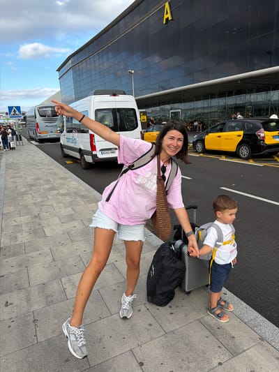 A woman and a young child stand on a sidewalk in front of an airport building. The woman, on the left, wears a pink t-shirt, denim shorts, and gray sneakers. She has a brown woven bag slung over her shoulder and is pointing with her left hand. The child, on the right, wears a white t-shirt, blue shorts, and gray sandals, with a yellow and gray backpack. The child is holding the woman's hand. They are next to a silver suitcase and a black duffel bag. In the background, an airport terminal with a large yellow "A" sign is visible. Several buses and yellow taxis are parked along the road. The ground is paved with gray concrete tiles. The weather appears to be cloudy with some sunlight. The location is identified as the city of El Prat De Llobregat, Spain, specifically near the airport.