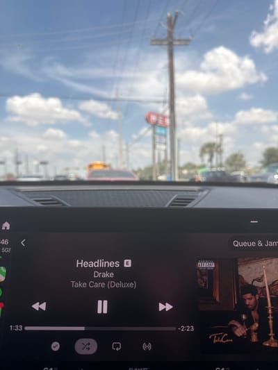 The image captures a view from inside a vehicle, looking forward through the windshield, with an infotainment system prominent in the foreground and a blurred urban street scene beyond.
The infotainment screen displays a music player interface. The track "Headlines" by Drake, from the album "Take Care (Deluxe)," is shown as playing but paused at the 1:33 mark, with 2:23 remaining. The album art, visible on the right, depicts a blurred image of Drake with a "PARENTAL ADVISORY EXPLICIT CONTENT" label, seated at a table with a goblet and a candlestick, accompanied by the album title "Take Care." Standard playback controls (previous, play/pause, next), shuffle, and repeat icons are present. The screen also shows "46" and "5G" indicators in the top-left, and a "Queue & Jam" button in the top-right. A dark, textured dashboard section with a speaker grille is visible directly above the screen.
The background, rendered out of focus, depicts a daytime environment under a blue sky with scattered white cumulus clouds. A prominent wooden utility pole with multiple crossarms and power lines is visible. Behind it, a red and white commercial sign, with letters "ES" partially visible, indicates a retail or service establishment. A multi-lane road is populated with several blurred vehicles, including a red car in the mid-ground and a yellow vehicle further to the right. Distant trees, including palm trees, are visible along the horizon. This scene is situated within the city of McAllen, United States of America.
