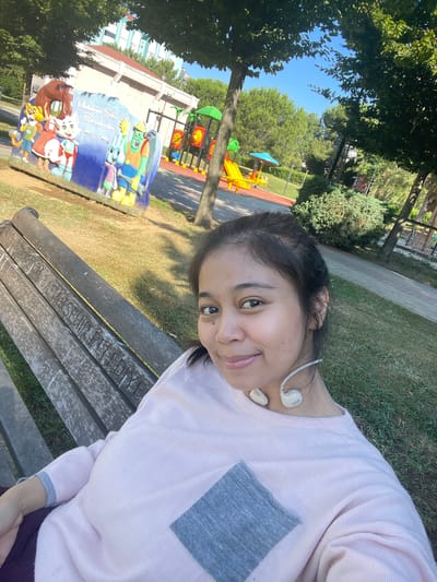 A young woman with dark hair pulled back and wearing a pale pink sweater with a gray square patch on the chest sits on a wooden park bench in Başakşehir, Turkey. She is smiling and looking at the camera. White earbuds are draped around her neck. The bench is weathered and has carved lettering that appears to spell "BAŞAKŞEHİR BELEDİYESİ," which translates to Başakşehir Municipality. In the background, a colorful mural depicting cartoon characters and a playground with slides and swings are visible. Lush green trees and grass surround the area. The sky is clear and blue.