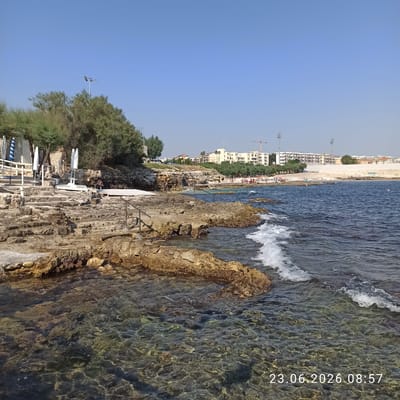 The image depicts a rocky coastline in Molfetta, Italy, on a clear, sunny day. The foreground features the shallow, clear water with visible rocks beneath the surface. The rocky shore is uneven and textured, with some areas appearing to be man-made steps leading down to the water, reinforced with metal railings in some sections. Further back, there are beach umbrellas and seating areas set up on a wooden platform, suggesting a recreational or beach club area. Vegetation, including trees and bushes, grows along the top of the rocky embankment. In the background, a city skyline is visible, characterized by multiple buildings, some of which appear to be apartment complexes or hotels, along with some construction cranes. The sea is a deep blue, with small waves breaking against the rocks. A few people are scattered on the distant sandy beach and in the water, indicating a public beach area. A date and time stamp, "23.06.2026 08:57", is visible in the lower right corner of the image.