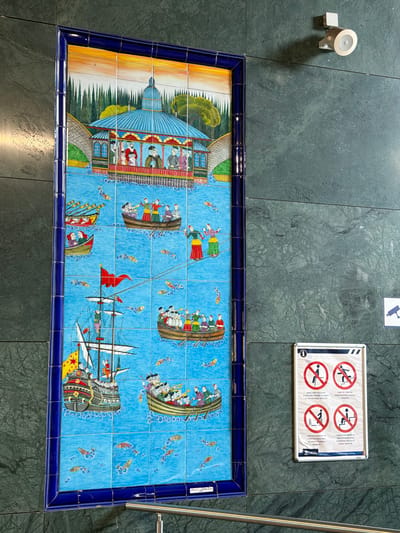 The image depicts a large ceramic tile mural depicting a historical scene set on water. The mural features several boats, including a sailing ship with multiple masts and flags, and various smaller rowboats carrying passengers. Figures in traditional attire are depicted in the boats and on a structure resembling a pavilion or building with a domed roof in the background. The background also shows a stylized landscape with cypress trees and a vibrant orange sky. Below the mural, a metal railing is visible. To the right of the mural, on the dark green marbled wall, is a white informational sign with several red-circled icons indicating prohibitions related to sitting and stair usage, along with text in Turkish. A security camera is mounted above this sign. The overall environment appears to be an indoor public space, possibly a transportation hub or cultural venue in Eyüpsultan, Turkey, given the Turkish signage.