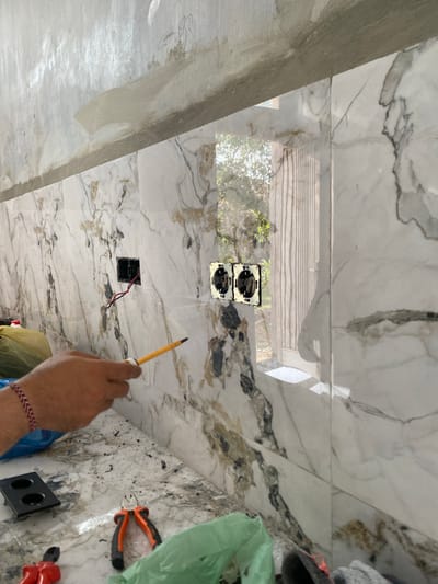 The image depicts an indoor scene, likely a construction or renovation project, given the exposed electrical outlets and wiring. The wall is covered in large, glossy marble-patterned tiles, predominantly white with black and gold veins. In the foreground, a person's right hand, adorned with a braided red and white bracelet, holds a yellow pencil or stylus. The hand is positioned near an unfinished electrical box with red wires emerging. Above and to the left of this, two completed electrical outlets are visible, mounted on the tiled wall.
Further to the right, a pair of orange and black pliers and a red electrical tool are laid out on the marble surface, alongside a black electrical box with two openings. A green plastic bag is also present in the background on the right. The lighting suggests an indoor setting, possibly during daylight hours, as evidenced by the light filtering through what appears to be blinds or louvers visible in the reflection on the wall tiles. The overall impression is one of ongoing electrical work within a tiled interior space.