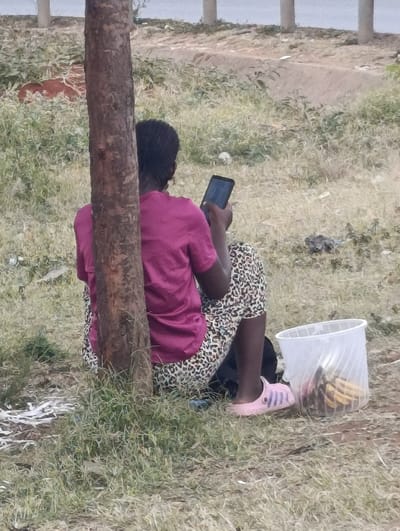 The scene, located in Matungulu west ward, Kenya, depicts an individual seated on sparse, dry grassy ground, with their back to the viewer and partially obscured by a tree trunk positioned centrally. The person wears a maroon short-sleeved shirt, leopard-print trousers, and pink sandals with a blue stripe. They hold a black smartphone in both hands, actively viewing its screen. To the individual's right, a translucent white plastic bucket contains several yellow bananas. A dark bag or object is partially visible behind their legs, near the tree base. The immediate ground consists of dirt and dry, sparse grass, with scattered white debris visible to the left. In the background, beyond a raised dirt bank or path, several light-colored vertical structures are discernible against a blurry, light grey expanse, possibly suggesting water or a distant paved area.