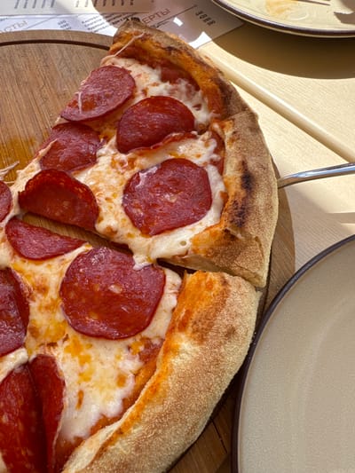 A close-up, top-down view of a pepperoni pizza slice on a wooden board is presented, with a portion of a beige plate and a metal serving utensil visible in the foreground and right side of the image, respectively. The pizza slice features a thick, golden-brown crust generously topped with melted mozzarella cheese and numerous slices of pepperoni. The background includes a blurred menu with Cyrillic text and prices, suggesting a restaurant setting. The scene is illuminated by natural light, casting soft shadows. The location is identified as Abrau-Dyurso, Russia.