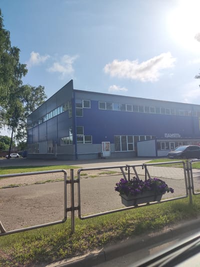 The image shows a blue and gray modern building with large windows, possibly a business or industrial facility. A sign with the Russian word "ПАМЯТЬ" (Memory) is visible on the building. In front of the building, there is a paved area with a road and a metal fence. A planter with purple flowers is attached to the fence. A white car is parked on the left side of the image, and a black car is parked in front of the building on the right. Trees are visible on the left and right edges of the frame. The sky is blue with some scattered clouds, and the sun is shining brightly, causing a lens flare in the upper right corner. The overall scene appears to be an exterior shot of a commercial property on a sunny day. It's not possible to determine the specific location within Polatsk, Belarus from the image alone.