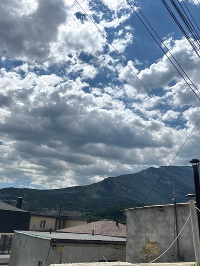 A partially clouded sky hangs over a hillside landscape. In the foreground, rooftops of several buildings are visible, constructed from materials including dark asphalt shingles, brown shingles, and light grey concrete. Some buildings appear to be residential or commercial structures. A tall, dark chimney extends upwards from one of the buildings. Further back, a green, tree-covered mountain range dominates the middle ground, with a rocky peak visible towards the top. The sky above is a mix of bright blue patches and white and grey cumulus clouds, with several dark power lines stretching diagonally across the upper portion of the frame. The scene suggests a residential area at the base of a mountain in a natural environment, potentially within the city of Yalta, Ukraine, given the geographic context. No people are visible in the image.