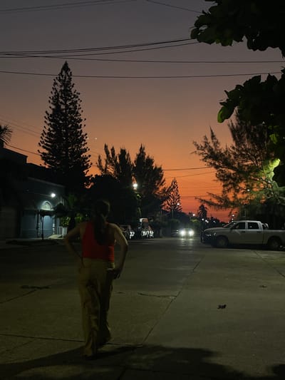 The image depicts a twilight street scene in Coatzacoalcos, Mexico. The sky transitions from a deep orange at the horizon to a dark purple-gray overhead, with two distinct celestial bodies, likely Venus and a crescent moon, visible above the silhouetted trees. Numerous overhead power lines traverse the upper frame.
In the foreground, a single individual stands with their back to the camera, positioned on the paved street. The person has dark hair pulled back into a ponytail and is wearing a red sleeveless top and light-colored, loose-fitting pants. Their hands are on their hips, and a long shadow extends from their body onto the ground.
The street, appearing to be asphalt or concrete with visible cracks, extends into the distance. On the left side, a dark blue/gray building with arched openings is visible, illuminated by a street light. Several tall, coniferous trees and other deciduous trees line both sides of the street, silhouetted against the vibrant sky. Multiple streetlights are active, casting localized illumination.
Vehicles are present in the mid-ground: a white pickup truck (resembling a RAM) is parked on the right side of the street, facing away from the camera. Further down the street, a vehicle approaches, indicated by its illuminated headlights creating a bright glare. Other parked cars are faintly visible on the left side of the street, obscured by shadow and distance.