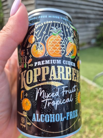 A person's hand with a light pink manicure is holding a black can of Kopparberg Mixed Fruit Tropical alcohol-free cider. The can is covered in condensation droplets. The label on the can features a gold pineapple, oranges, and other tropical fruit illustrations, as well as the words "PREMIUM CIDER," "KOPPARBERG," "Mixed Fruit Tropical," and "ALCOHOL-FREE" in gold and blue lettering. The background is blurred and appears to be an outdoor setting with green grass and a wooden fence, consistent with a garden in West Suffolk, United Kingdom.