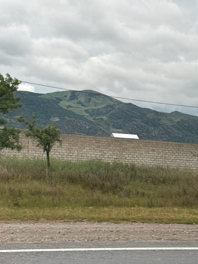 This daytime landscape image, taken in Korkmaskala, Russia, depicts a serene, overcast scene. A vast mountain range, with slopes exhibiting shades of dark green and grey, dominates the background, rising under a soft, cloudy sky. Thin, dark lines, likely power cables, are visible cutting across the upper sky and parts of the mountain.

In the foreground, on the right side, a paved road with a clear white line along its edge extends horizontally. Adjacent to the road, a strip of dry, light-brown earth transitions into a wider area of green grass and varied vegetation. A light-colored brick wall runs horizontally through the mid-ground, acting as a clear division between the grassy area and the mountainous terrain. Positioned on or near this wall, against the dark mountain, is a small, bright white rectangular object, possibly a sign or a simple structure.

Several trees are scattered throughout the scene; one prominent tree with green foliage stands near the brick wall and the mountain, while others are partially visible on the left. The sky is completely covered by thick, diffused clouds, indicating an overcast day without direct sunlight. No people or specific activities are present, and there is no visible text. The overall impression is a calm, rural or semi-rural environment.