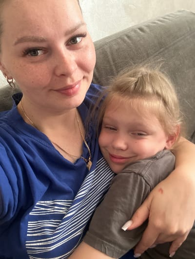 A close-up, indoor selfie shows a woman with blonde hair pulled back, wearing a blue t-shirt and a gold necklace with a cross pendant. She is smiling and has her arm around a young blonde child who is resting their head on the woman's shoulder and also smiling. The child is wearing a dark gray t-shirt. They are both on a gray sofa. The image was taken in Abakan, Russia.