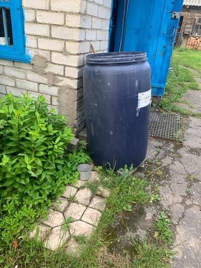 The image shows an outdoor scene in Відзаўскі сельскі Савет, Belarus. In the foreground, a large, dark blue plastic barrel stands next to a weathered brick building with a blue wooden door. Lush green foliage surrounds the barrel and a section of paved ground. A small metal cup is visible amongst the plants. A metal grate is partially visible near the barrel. The building appears to be a functional structure, possibly a shed or outbuilding, with a visible window on the left side. The overall impression is one of a rural or semi-rural setting, with evidence of recent construction or repair work on the building, indicated by the patch of wet cement on the brick wall.