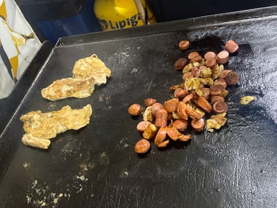 The image shows a flat-top grill with food being cooked. On the left side of the grill are two pieces of what appears to be chicken or fish, cooked to a golden brown. To the right of the chicken/fish are numerous sliced pieces of sausage and some fried potato chunks. The grill surface is dark and appears to be well-used, with some oil or grease present. In the background, out of focus, is a yellow propane tank with some blue and black objects. The context of Iquique, Chile, is not directly visible in the image itself.