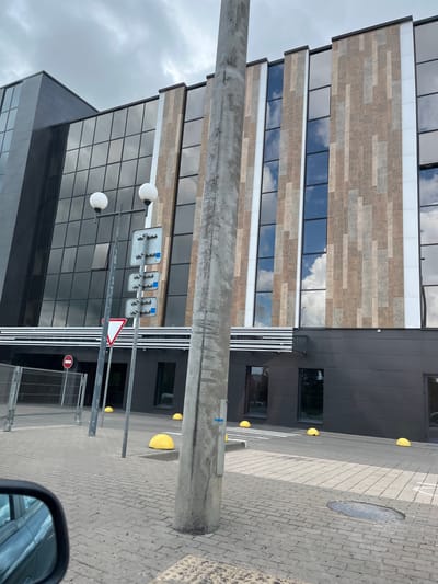 The image depicts the exterior of a modern building with a dark grey facade and a section featuring a pattern of vertical wooden-like tiles. The building has large, dark windows reflecting the cloudy sky and trees. In front of the building, there is a paved area with yellow semi-spherical bollards and a metal fence. A concrete pole with traffic signs is prominently positioned in the foreground, obscuring part of the building's facade. The scene appears to be a street or public area in a city. The location is identified as Папярнянскі сельскі Савет, Belarus.