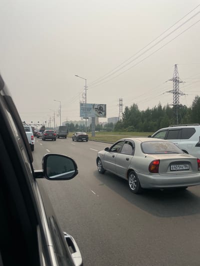 This is a photograph taken from inside a car, looking out at a traffic jam on a hazy day. The road is paved and appears to be a multi-lane highway. Several cars are visible in the distance, all stopped or moving very slowly. The sky is overcast and a light gray, suggesting either smog or fog. Tall electrical pylons with multiple wires are prominent in the background, a common sight in industrial or power-line corridors. Trees line the side of the road, partially obscured by the hazy atmosphere. In the foreground, parts of the car the photo is taken from are visible, including a side mirror and the car's roofline.
One car in the middle ground, a silver sedan, has the license plate E401PK 186 RUS. There is also a white SUV behind it. Further down the road, a billboard with Cyrillic text is visible, partially obscured by traffic. The overall impression is one of a standstill on a highway, possibly due to poor visibility or a traffic incident. The time of day appears to be daytime, given the lighting. The location is identified as Surgut, Russia.