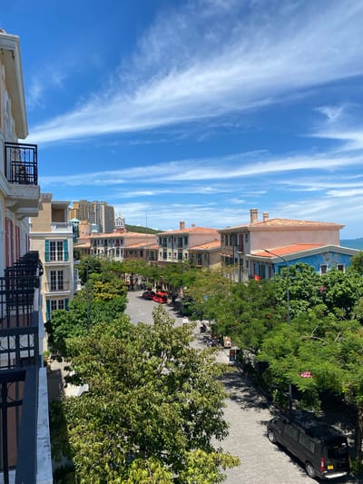 The image depicts a view of a street lined with buildings, possibly in a resort town, under a bright blue sky with wispy white clouds. The perspective is from a higher vantage point, looking down onto the street and the facades of the buildings.

The buildings feature a variety of architectural styles, with some having terracotta roofs and ornate balconies, suggesting a Mediterranean influence. Others are more modern, with a cluster of tall buildings visible in the background. Lush green trees are abundant, framing the street and casting shadows on the paved road.

On the street, there are several objects, including a black van with the word "LUXURY" printed on its side. A red vehicle is also parked on the street. A few motorcycles are visible, suggesting local transportation.

The time of day appears to be daytime, with the sun casting distinct shadows. The weather seems to be pleasant and clear. While there are no people clearly visible in the image, the presence of vehicles and buildings implies human activity.

The overall scene evokes a sense of a vibrant, possibly tourist-oriented location, with a mix of residential and commercial structures set against a backdrop of natural greenery and a striking sky. The location context of Phú Quốc, Vietnam, aligns with the visual elements of lush vegetation and a coastal or island feel.