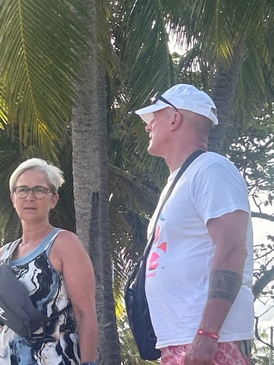 Two people stand near a palm tree in an outdoor setting. The background suggests a tropical location.

A man in a white t-shirt and white baseball cap stands to the right, his arm visible with a tattoo and a red bracelet. He is wearing a dark bag across his body and floral-patterned shorts. He appears to be looking down.

A woman with short, gray hair and glasses stands closer to the camera, wearing a patterned tank top and a dark bag across her body. 

The location may be Sainte-Anne, France, given the prompt. The scene likely depicts a casual moment during a vacation or outing.
