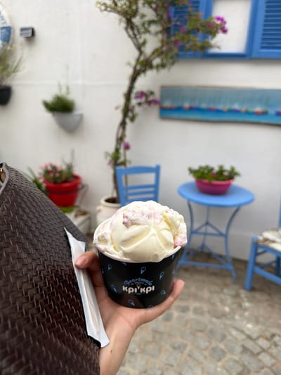 A person's hand holds a black paper cup filled with vanilla ice cream that has pink swirls. The cup has the word "kri kri" in white text, along with other small icons. The scene is outdoors in a courtyard with cobblestone paving. In the background, there is a white wall with blue shutters and decorative blue panels with figures. A small blue table with a pink pot of green plants sits nearby. A brown woven surface, possibly a chair or table, is in the foreground. The image appears to be taken during the daytime, with natural lighting. The overall setting suggests a charming, possibly Mediterranean, outdoor cafe or restaurant in Olympiada, Greece.