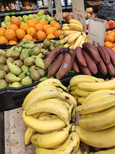This is a close-up, eye-level, outdoor photograph of a fruit stand in Navapolatsk, Belarus. The focus is on a variety of fruits. In the foreground, a large pile of yellow bananas dominates the lower half of the frame. Behind them, to the left, are some red bananas. To the left and behind the bananas, there is a crate filled with green pears. In the middle ground and background, there are crates of oranges and other citrus fruits, and a few apples. The fruits are arranged in plastic crates, suggesting a market or grocery store setting. There are price tags visible with Cyrillic text and prices. The lighting is bright and natural, indicating it might be daytime. There are no people visible in the frame.