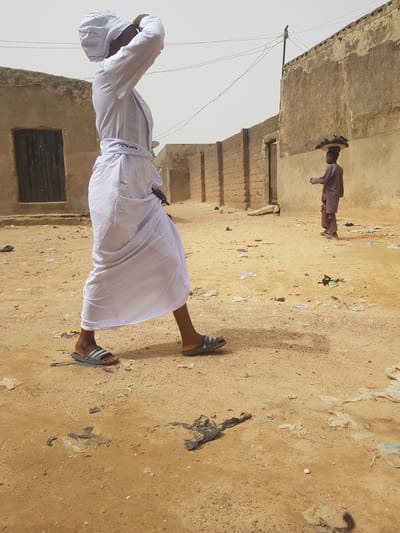 A medium shot captures two individuals in Dutsinma B, Nigeria. The foreground features a man facing away, clad in a white robe and head covering, standing on a dirt ground with scattered litter. He appears to be adjusting his head covering with both hands. In the background, another person in a long-sleeved maroon outfit and a straw hat, standing on the same dirt ground. The environment includes dilapidated structures on both sides, and the sky is overcast.