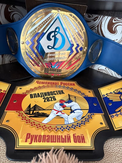 The image shows two championship belts. The top belt is blue with gold accents and features a stylized "D" logo and the text "ОБЩЕСТВО «ДИНАМО» 80 ПОБЕДА!" which translates to "Dynamo Society 80 Victories!". The belt also has text in Russian around the edge, including "ВСЕРОССИЙСКИЕ СОРЕВНОВАНИЯ ПО БОКСУ" (All-Russian Boxing Competitions) and "САНКТ-ПЕТЕРБУРГ 2023" (Saint Petersburg 2023). The bottom belt is gold and black with blue accents. It features an image of two people in a combat stance, a bridge, and the text "ЧЕМПИОНАТ РОССИИ ВЛАДИВОСТОК 2026" (Championship of Russia Vladivostok 2026) and "РУКОПАШНЫЙ БОЙ" (Hand-to-Hand Combat). The belts appear to be awards for martial arts and boxing competitions. The background suggests the belts are displayed indoors, possibly on a shelf or table. There is no indication or visual element that places this image in the city of Elovo, Russia.