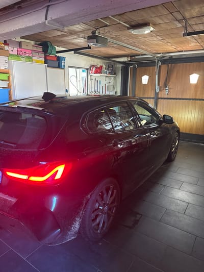The image displays a dark-colored BMW hatchback parked inside a residential garage located in Lingolsheim, France. The vehicle is positioned with its rear-left quarter visible, featuring illuminated red taillights with distinct L-shaped LED elements. The rear bumper and left rear wheel well show visible accumulation of dirt and splatter. The car is equipped with black multi-spoke alloy wheels and a black shark fin antenna on its roof.

The garage floor is covered with dark rectangular tiles, and a small wet patch is observable on the tiles beneath the vehicle's rear. The ceiling consists of exposed wooden panels, from which an automatic garage door opener mechanism and lighting fixtures are suspended. Along the left wall, a storage area includes white cabinets topped with various stacked boxes and storage containers. A window in this wall provides a reflective view of an exterior structure. The far end of the garage features a segmented wooden garage door, which incorporates two illuminated, truncated hexagonal window inserts. Shelving units holding various tools and objects are mounted on the wall section between the storage area and the garage door. No individuals are present in the scene, and no specific actions or interactions are depicted.