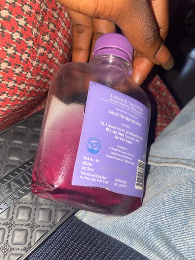 A person's hand, with dark skin and visible fingerprints, holds a clear glass bottle filled with a pink-purple liquid. The bottle has a purple cap and a purple label adhered to its side. The label contains text indicating it is "LACOCO LIQUEUR (VODKA & BERRY BLENDED DRINK)" and states it was "Manufactured in Nigeria By: SUNLIGHT FOOD INDUSTRIES LIMITED". The address listed is "No. 4 Laronpe Crescent, Opic/Fatgbem Bus Stop, KM 8 Lagos-Ibadan Expressway, Isheri, Ogun State, Nigeria." Ingredient information and production details like "Batch No.: 001", "Mfg Date: Exp: 12months", and an expiration date of "15/05/2026" are also present. The bottle is positioned on a textured, grey surface with a repeating pattern that resembles the Chanel logo, next to a blue denim fabric. In the background, a red and black patterned fabric is visible. The scene is lit, revealing the contents of the bottle and the label's text. The image does not provide any specific information about the location in the city of Barikin Gidar Kanat, Nigeria, as the address on the label indicates Ogun State.