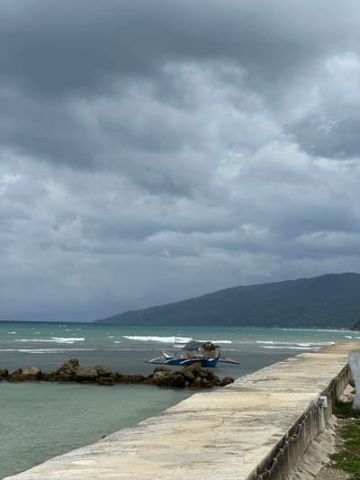 The image captures a coastal scene in Oslob, Philippines, characterized by a cloudy sky and a choppy sea. In the foreground, a concrete walkway stretches along the coastline, with visible electrical conduits running along its edge. To the left of the walkway, a body of water with greenish-blue hues laps against a cluster of large, dark rocks. Further out in the water, a small, blue outrigger boat with a thatched canopy is anchored. The sea displays turquoise and darker blue tones, with white waves breaking along the shore. In the background, a verdant, tree-covered mountain slopes down towards the sea. The sky is dominated by heavy, grey clouds, suggesting overcast or impending stormy weather. No people are visible in the frame.