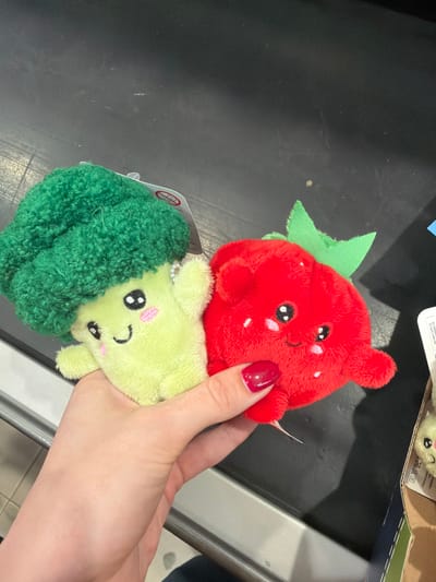 A person's hand holds two plush toys against a dark, possibly black, surface, likely a store counter or conveyor belt, in Berezniki, Russia. The hand, with red nail polish on the thumb and index finger, is fair-skinned. One plush toy is a bright red strawberry with a green leaf at its top. It has black eyes, a small smiling mouth, and pink blush marks on its cheeks, along with white embroidered 'seeds' on its body. The second plush toy is a light green, soft-textured broccoli head, with a darker green, more textured 'stem' or florets wrapping around it. Like the strawberry, it features black eyes, a small smiling mouth, and pink blush. Both toys appear to be part of a set of cute, anthropomorphic fruit and vegetable plushies.

In the background, to the upper right of the red strawberry, a white product label with black text and symbols is partially visible. Legible portions include "(01) 062020", "(21) 3JPS", and "Fraberik". A white tag with a red circle-slash symbol (often indicating a safety warning) is also partially visible near the green broccoli. The setting appears to be indoors, possibly a retail environment, given the presence of product labels and the type of counter. The overall impression is one of playful, soft toys being presented or purchased.
