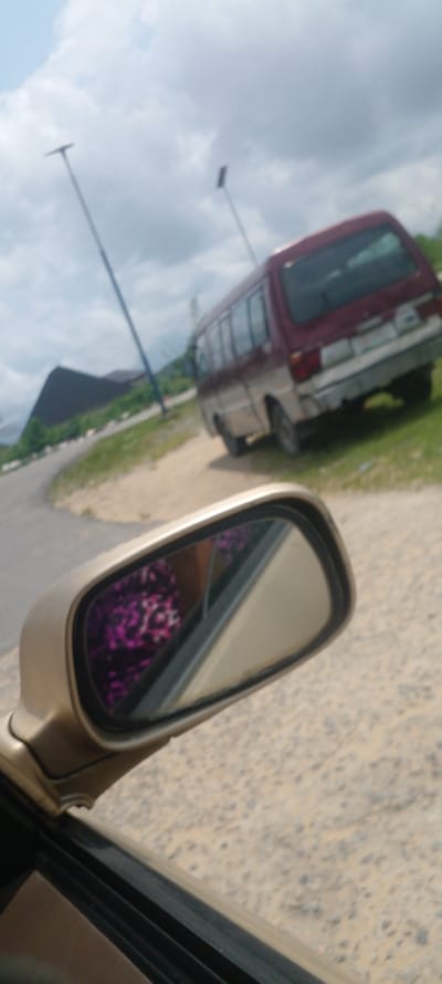 The image captures a scene from within a vehicle, focusing on the side-view mirror and the exterior. The mirror reflects a person wearing a brightly colored, patterned garment, likely with floral motifs in shades of pink and purple. Beyond the mirror, the scene shows a reddish-maroon and silver mini-van parked on the side of a dusty, unpaved road. The van appears to be in moderate condition with some signs of wear and tear on its rear bumper. To the left of the van, a dark, pyramid-shaped structure is visible against a cloudy sky. Two tall, slender poles, possibly streetlights or antenna masts, are also present in the background, one on the left and another further back. The environment suggests an outdoor setting in a somewhat rural or developing area within Yenagoa, Nigeria, with a mix of natural elements like grass and soil, and man-made structures. The road curves away into the distance, hinting at a journey or movement. The overall impression is of a casual observation captured during transit.
