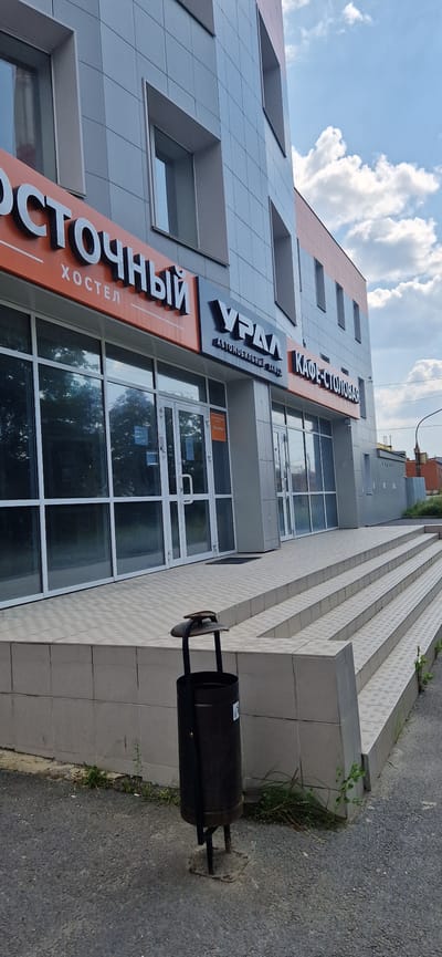 An exterior shot captures a modern building with a grey facade and orange and dark grey signage. The signs read "ОСТОЧНЫЙ ХОСТЕЛ" (Eastern Hostel), "УРАЛ" (Ural) with "АВТОМОБИЛЬНЫЙ ЗАВОД" (Automotive Plant) underneath, and "КАФЕ-СТОЛОВАЯ" (Cafe-Canteen). The building has large glass windows and doors, and a set of tiled steps leads up to the entrance. A dark-colored trash can stands to the left of the steps. The ground in front of the building is asphalt with scattered small stones. The sky above is blue with fluffy white clouds. In the background, a street lamp and some buildings are visible. This image appears to be taken in the city of Miass, Russia, as suggested by the "УРАЛ" signage which is associated with the Ural Automotive Plant, a major employer in Miass.