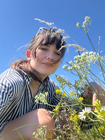 A low-angle, medium shot captures a woman in a field of wildflowers under a bright blue sky. The woman, with dark hair pulled back and bangs framing her face, looks directly at the camera with a slight smile. She is wearing a black and white striped button-down shirt and appears to be kneeling or crouching amidst the foliage. Her right hand is visible, gently touching some of the wildflowers, which include yellow blossoms and white daisies. The background is a clear, cloudless blue sky, with a few small dark specks that could be birds or insects visible in the upper left quadrant. The lighting suggests it is daytime, likely midday or early afternoon, casting shadows on the woman's face and leg. The overall impression is a serene moment in nature. The location is identified as Krasnyi Yar, Russia.