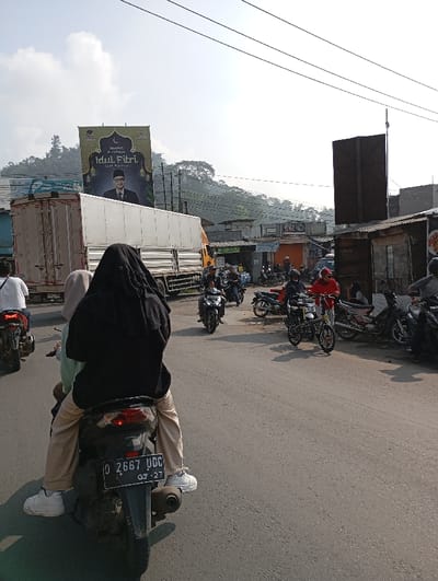 The image shows a street scene in Kutawaringin, Indonesia, with a billboard in the background. The billboard reads "Selamat Hari Raya Idul Fitri 1443 H" and features an image of a man in traditional clothing. In the foreground, there are several people riding motorbikes. There are also several motorbikes parked on the side of the road. The road is paved and there are power lines overhead. There are several buildings in the background, including a large building with a flat roof, a smaller building with a corrugated metal roof, and a building with a wooden facade. The image is taken during the day with clear skies. The scene is a typical street scene in Kutawaringin, Indonesia.