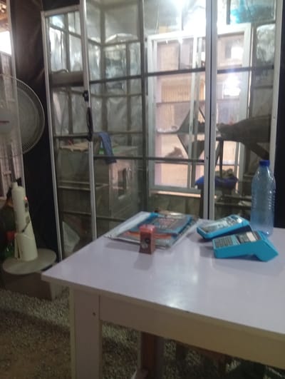 The image captures an indoor or semi-enclosed commercial setting, likely a shop or small business in Barako, Nigeria. No people are visible in the frame.
In the foreground, a light-colored rectangular table occupies the lower right portion of the image. On the table, several objects are arranged: two blue point-of-sale (POS) terminals or card readers, a clear plastic water bottle that is partially filled, a stack of books or magazines (one with a red cover, another blue with the word "MATHS" partially visible), and a small, reddish-brown rectangular object.
To the left of the table, a white standing fan with a circular protective cage is positioned. Its cream-colored base shows indicator lights, one red and one green, and part of a brand name, "Binatone", is faintly discernible.
The background is dominated by a large, multi-paned structure with an aluminum frame, resembling a display case or a room divider made of glass or clear plastic. Through this structure, various items are vaguely visible, including what appears to be a silver-colored oven or industrial equipment with a handle. A blue cloth or towel is draped inside this enclosure. Beyond this, faint outlines of what might be windows or another structural element are visible, suggesting an area with more light. The flooring in the foreground and under the fan appears to be gravel or coarse aggregate.
The presence of POS terminals suggests the setting is a place where transactions occur. The standing fan indicates a warm climate, consistent with the location context. The overall impression is that of a functional, operational business environment. The time of day appears to be daylight hours, given the ambient illumination.