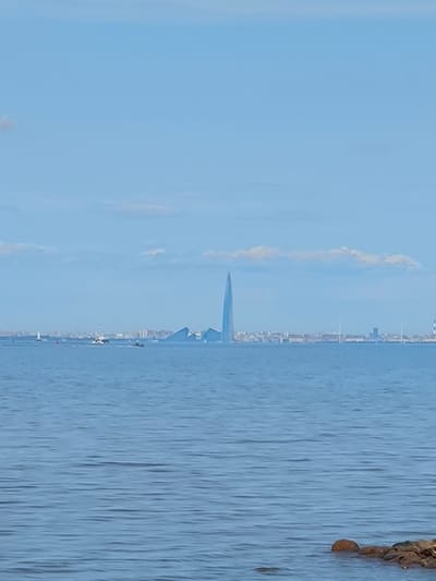 The media file shows a wide shot of a body of water, likely the Gulf of Finland, under a bright, hazy sky with scattered white clouds. In the distance, a modern cityscape is visible, dominated by a uniquely shaped, spiraling skyscraper that appears to be the Lakhta Center in St. Petersburg. The skyscraper is a prominent architectural feature of the scene.

Several small boats are scattered across the water, indicating recreational activity or perhaps local transport. A lighthouse is also visible on the left side of the frame, suggesting a coastal or navigational area. The water is calm with gentle ripples, reflecting the light of what appears to be midday. The overall mood is serene and expansive, with the vastness of the water and sky emphasizing the scale of the distant urban development. The color palette is dominated by blues and grays, with touches of white in the clouds and subtle hues of the distant buildings. The image does not contain any visible text. The scene is likely captured from a shore or a vessel, looking towards the horizon and the city.