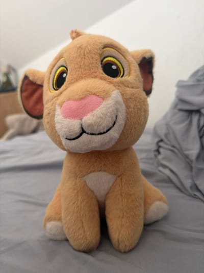 The image shows a close-up of a plush toy lion cub sitting on a gray surface, which appears to be a bed. The toy is golden brown with large yellow eyes outlined in black and yellow, a pink nose, and a white patch on its chest and front legs. The background is blurred but suggests an indoor environment with a white wall and a gray blanket or comforter. The location of this scene in Falenty Nowe, Poland, is not discernible from the image itself.