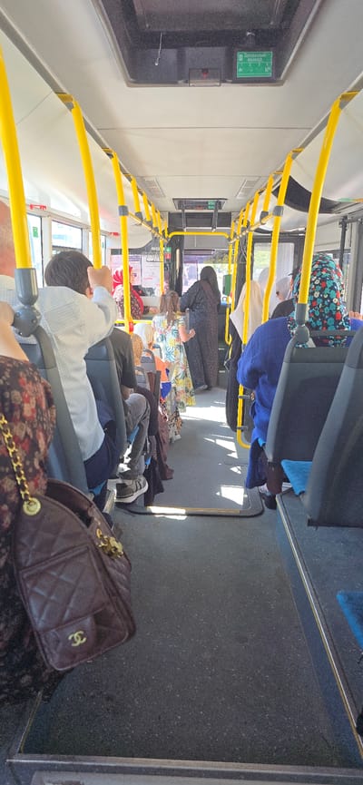 The image shows the interior of a bus in Grozny, Russia. The bus is populated with several passengers, some of whom are wearing hijabs, which are common in the region. The overall environment is a typical public bus interior, with yellow grab poles and rows of seats. The lighting suggests it is daytime, with sunlight streaming through the windows, casting shadows on the floor. Some passengers are seated, while others appear to be standing or moving. The focus is on the general scene inside the bus, with no particular individual or action being the primary subject. The bus appears to be in motion or stopped at a station.