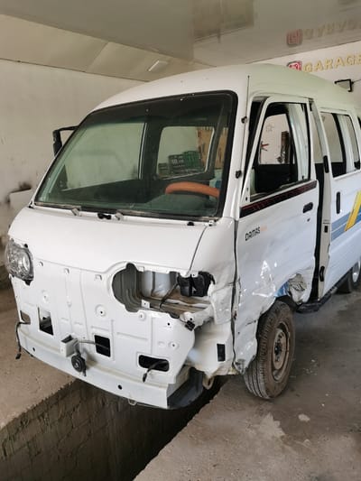 In a garage in Karabair, Uzbekistan, a white Daewoo Damas van with significant front-end damage is parked. The hood, front bumper, and right fender have been removed or are severely damaged, exposing internal components. The right side of the van shows extensive crumpling from a collision. The vehicle is a light commercial van, typically used for cargo or passenger transport, and its current state indicates it is undergoing repairs or salvage. The garage environment appears functional but unrefined, with exposed brickwork and concrete flooring. Tools and equipment are visible in the background, suggesting ongoing work. The lighting is artificial, typical of an indoor workspace. The overall scene is one of mechanical repair and vehicle damage.