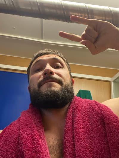 A close-up selfie shows a man with a beard wearing a pink towel. His head is tilted slightly, and his eyes are looking towards the camera. Above his head, a hand is making the "rock on" gesture. The background appears to be indoors, possibly a locker room or bathroom, with a large metallic pipe running along the top of the frame and a blue wall to the left. The overall lighting is warm and slightly dim.
