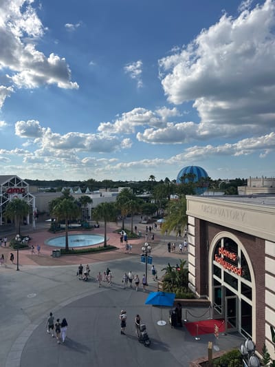 This is an aerial view of a public area at Disney Springs in Lake Buena Vista, Florida, United States. On the right is Planet Hollywood Observatory, a restaurant building with a white "OBSERVATORY" sign on top of it. People are walking on the street in front of the building. A large blue and white striped balloon is visible in the distance. The sky is blue with scattered white clouds. There are palm trees lining the streets. The AMC cinema building is visible in the bottom left corner. In the middle is a circular water fountain and lampposts.