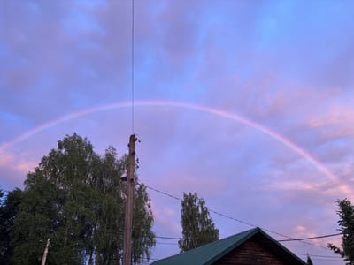 A rainbow arches across a dusky, purple and pink sky in Zaprudnya, Russia. A tall, weathered wooden utility pole with wires and a lamp stands to the right of center. To the left and partially in front of the pole, a dense cluster of green trees is visible. In the lower right corner, the dark green, angled roof of a wooden house juts into the frame. The sky is filled with soft, diffused clouds, and the overall impression is one of twilight serenity.