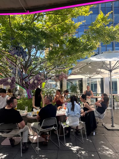 People are seated at a long table on a rooftop or patio. The table is decorated with flowers and lights. Green trees overhang the table, providing shade. Above them, there is a building and patio umbrellas. Individuals are conversing while seated on white metal chairs at the tables. The scene is bathed in natural light.
