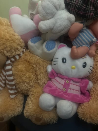 The image shows a pile of stuffed animals. In the foreground is a white Hello Kitty plush toy wearing a pink dress. To the left is a large tan teddy bear with a striped scarf. Partially visible are other stuffed animals including a white bunny and a doll wearing blue and white striped overalls. The background is blurred. The scene is indoors, with the stuffed animals arranged on a dark surface. There is no indication that this image is located in San Antonio, Venezuela, or any specific geographical location.