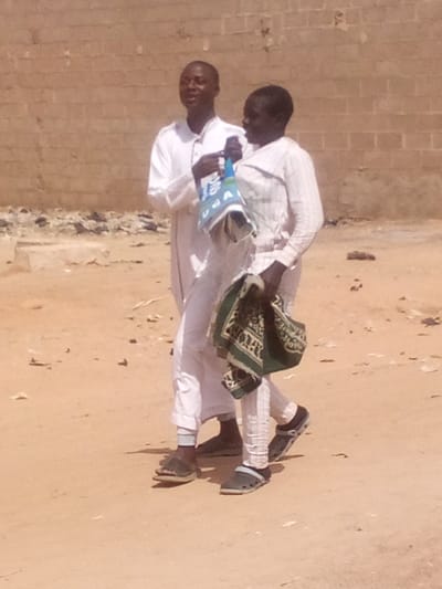 Two individuals, appearing male, walk across a sandy or dirt ground in Rangaza, Nigeria. The individual on the left wears a white, long-sleeved tunic and dark sandals, holding a blue and white patterned bag with "UDA" visible text. Their head is turned towards the second individual, mouth slightly open. The individual on the right wears a light-colored, possibly striped, long-sleeved top and matching trousers, with dark clogs featuring a lighter strap. This individual carries a folded green and beige patterned fabric in their left hand and looks towards the first individual, also with their mouth slightly open. In the background, a light brown brick wall is visible, with some scattered debris at its base. Bright, natural sunlight illuminates the scene, casting minimal shadows.