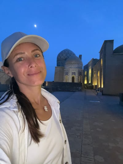 This is a portrait-oriented, vertically-aligned selfie taken outdoors at twilight, likely in Samarkand, Uzbekistan, given the architectural style. The main subject is a woman with fair skin, blue eyes, and dark brown hair, wearing a white baseball cap and a white shirt or jacket. She is looking directly at the camera with a slight smile, appearing relaxed and content.
Behind her, to the left, is a section of a historical building with intricate tilework on a dome, characteristic of Central Asian Islamic architecture. The building is illuminated by warm, artificial lights, suggesting it is evening. To the right of the dome, a larger structure with multiple arched windows, also lit from within, stretches into the background. The sky is a deep, clear blue, indicating the transition from day to night, with a small, bright crescent moon visible in the upper left quadrant of the image. The ground appears to be paved.
The overall mood is serene and peaceful, amplified by the soft lighting and the twilight setting. The woman's attire, particularly the white cap and shirt, is simple, allowing the background architecture and the natural light to be prominent. The photo captures a moment of enjoyment while exploring a historically significant location at a beautiful time of day.