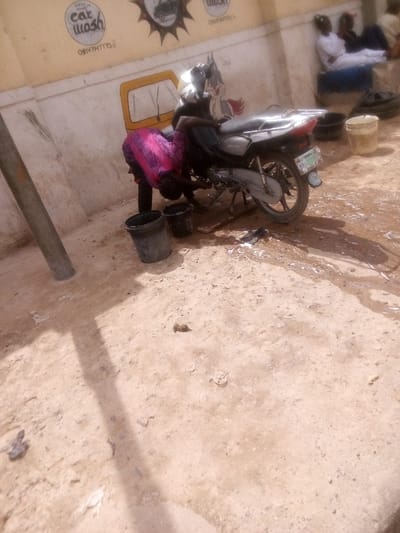 The image displays an outdoor scene in Tudun Murtala, Nigeria, depicting a motorcycle wash operation. In the foreground, the ground consists of light-colored, dry earth, interspersed with wet patches and soapy runoff originating from the washing activity. A grey, cylindrical pole is vertically positioned on the left side.
A single individual, dressed in a bright pink patterned top over dark trousers, is actively washing a silver-grey motorcycle positioned on its kickstand. This person is bent over, focused on cleaning the lower engine and chassis components of the vehicle. Two dark-colored plastic buckets are present on the ground beside the motorcycle, one containing water or soap solution. A dark rag or sponge lies on the wet ground near the motorcycle's rear wheel, which bears a visible Nigerian license plate.
In the background, a light yellow building wall serves as the backdrop. It is decorated with painted graffiti, including a circular "Car Wash" logo, several phone numbers (one clearly visible as "08147747775"), a cartoon character resembling Bugs Bunny, and a yellow outline of a vehicle. To the right, three additional individuals are observed seated or reclining against the wall; two are attired in light-colored, possibly white, traditional garments. They appear to be observing the washing activity.