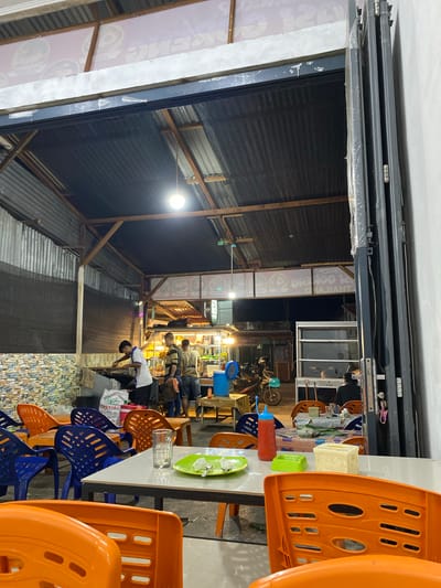 The image shows an outdoor seating area of a restaurant in Leuhan, Indonesia. The scene is viewed from the perspective of someone seated at a table. In the foreground, there are bright orange plastic chairs. A white table is set with a green plate containing food and cutlery, a glass, a red squeeze bottle, a green ashtray, and a small white box. Behind the table, blue and orange plastic chairs are arranged around other tables, some of which are covered with patterned cloths.
In the middle ground, there is an open-air food stall or kitchen area where three men are visible. One man in a white shirt is standing behind a counter and appears to be cooking or preparing food. Two other men, one in a striped shirt and the other in a lighter colored shirt, are standing nearby. A motorcycle is parked to the left of this area, with a blue cooler bucket on a nearby table.
Further back, the roof of the establishment is visible, constructed with corrugated metal and wooden beams. A sign with text in a foreign language, likely Indonesian, is mounted above the food stall. The background shows a dimly lit street scene, with glimpses of other buildings and a parked vehicle. The overall environment appears to be a casual, open-air dining establishment.