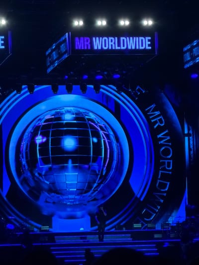 A man stands on a stage in front of a large, circular, blue LED screen displaying a globe. Above the screen, illuminated signs read "MR WORLDWIDE" and "PULL-PITBULL-PITBULL". The stage is lit with blue lights, and there is a band setup on the right side of the stage. The image is taken from an audience perspective. The location is not specified in the image, therefore the city of Lija, Malta cannot be determined.