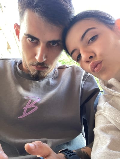 The image shows a close-up selfie of a young man and woman, likely a couple, taken outdoors. The man, on the left, has dark hair, a beard, and is wearing a grey sweatshirt with a purple "B" logo. He is looking directly at the camera with a serious expression. The woman, on the right, has dark hair and is puckering her lips for a kiss. She is wearing a light-colored hooded sweatshirt. They are positioned closely together, with the woman's head resting on the man's. The background is out of focus, but appears to be an outdoor setting with greenery, possibly a park or garden. The lighting suggests it is daytime. The image is a selfie, implying they are taking the picture themselves. The overall impression is one of intimacy and affection. The specific location in the city of Karlovo, Bulgaria cannot be determined from the image alone.