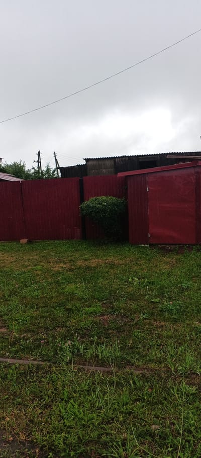 The image shows a residential backyard in Podgornoye, Russia, on a cloudy day. A dark red wooden fence runs across the middle ground, separating a grassy lawn from structures behind it. To the left, the fence is solid and unbroken. In the center, a small, dense green bush is visible in front of a section of the fence. To the right of the bush, there is a red shed or small outbuilding with a visible door. Behind the fence and shed, the tops of some buildings with corrugated roofs are visible, along with utility poles and wires. The foreground is a grassy area with various types of grass and some small wildflowers. A narrow, dark, raised strip, possibly a curb or border, runs horizontally across the lower part of the frame. The sky is overcast and a light gray.